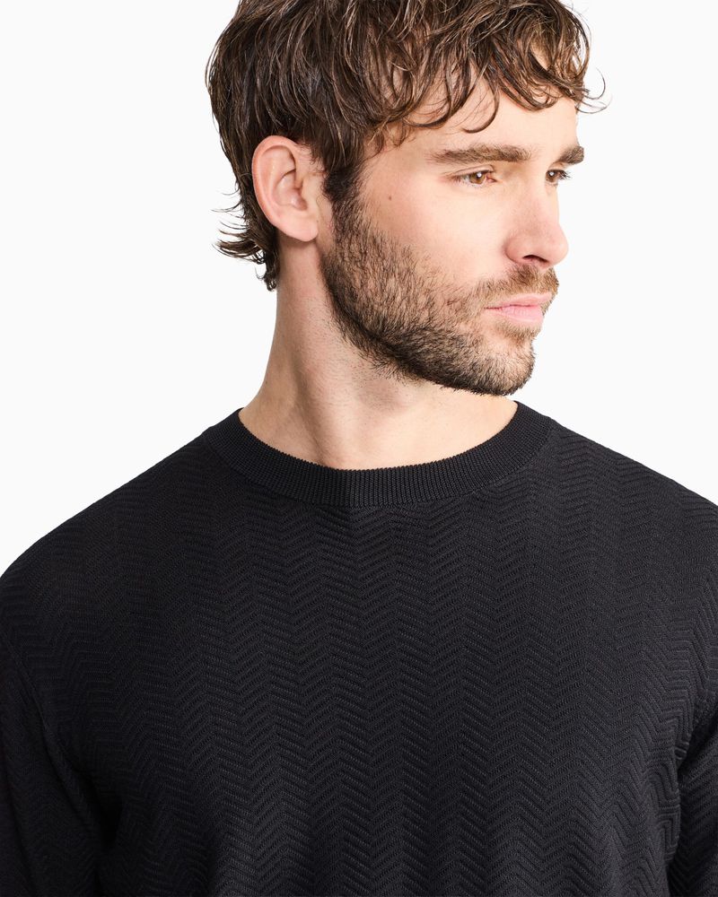 Close-up of a man with light brown hair and beard wearing a black textured knit crewneck sweater against a white background.