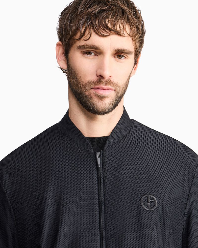Portrait of a man wearing a black textured zip-up bomber jacket with a round chest logo against a white seamless background.