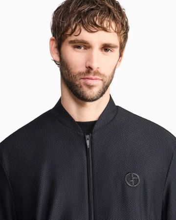Portrait of a man wearing a black textured zip-up bomber jacket with a round chest logo against a white seamless background.