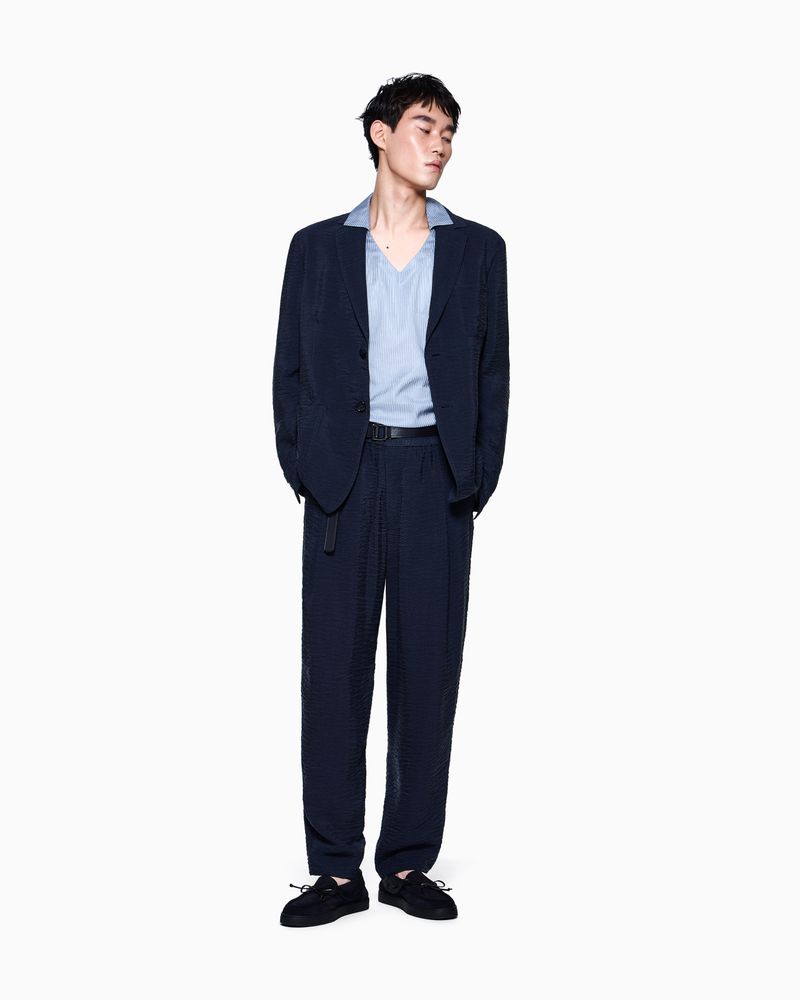 Model wearing a navy blue textured suit and light blue collar shirt standing against a seamless white studio background.