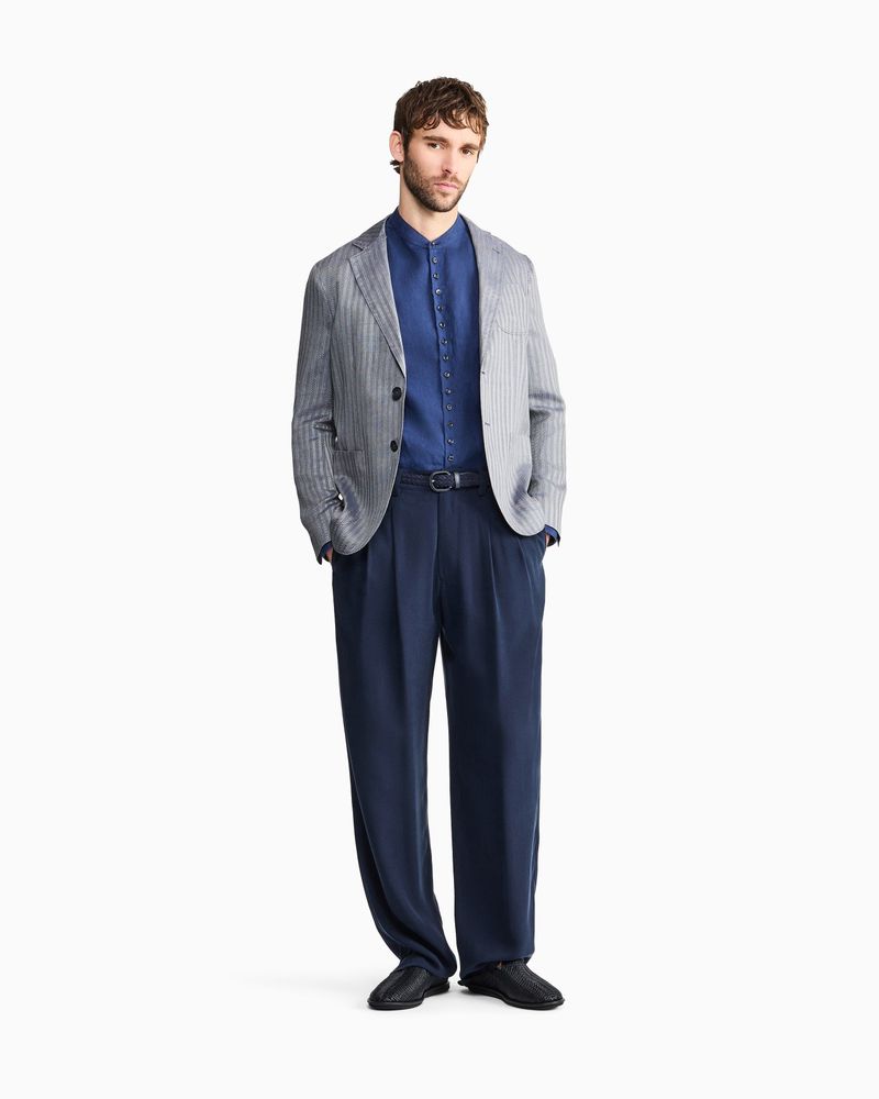 Model posing in a grey pinstripe blazer, blue shirt, and navy pleated trousers against a seamless white studio background.
