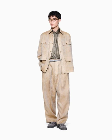 East Asian male model wearing glasses, a tan field jacket, and pleated trousers against a white seamless backdrop.