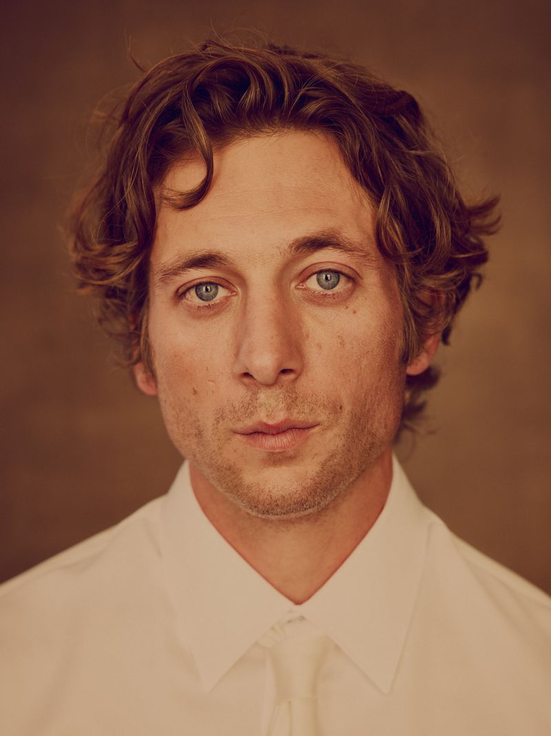 Close-up portrait of actor Jeremy Allen White wearing a white collared shirt and tie against a warm brown studio backdrop.