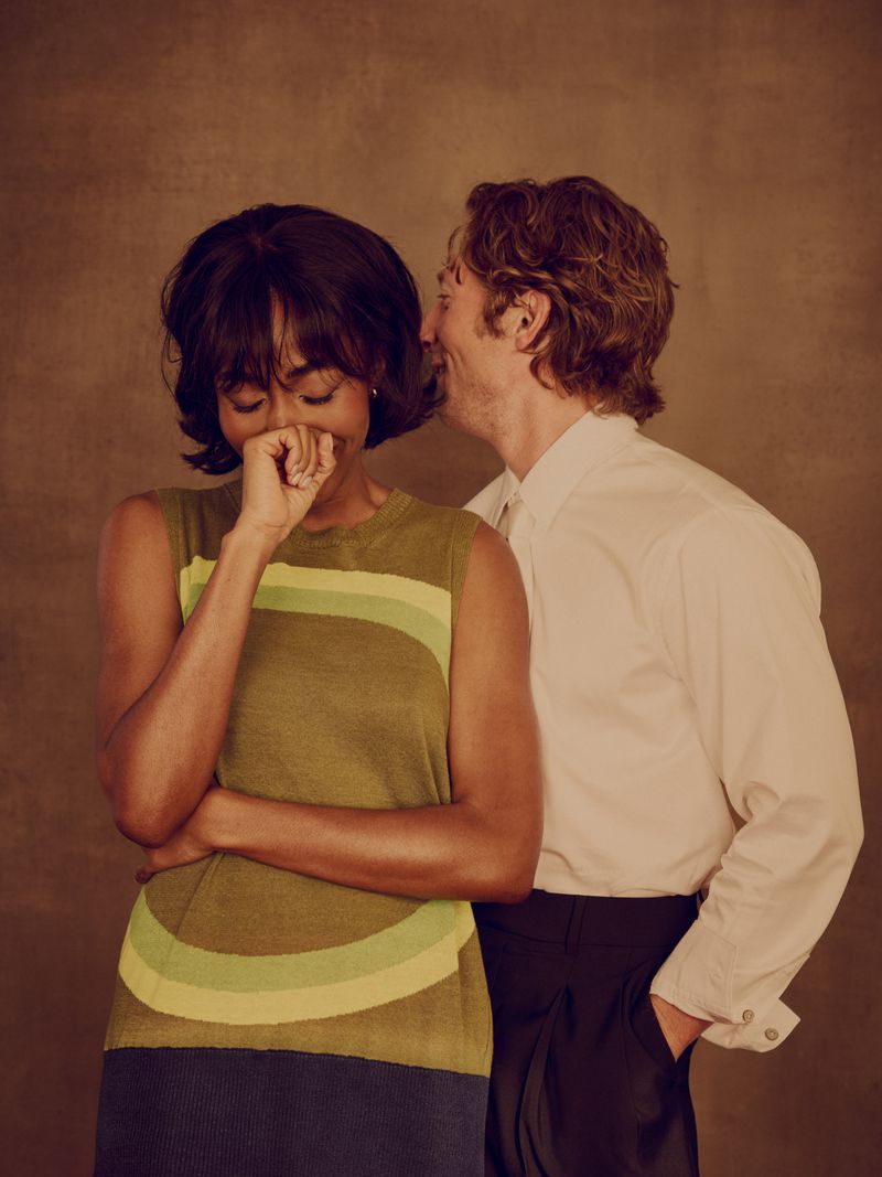 Ayo Edebiri laughing in a green patterned knit top beside Jeremy Allen White in a white shirt against a warm brown backdrop.