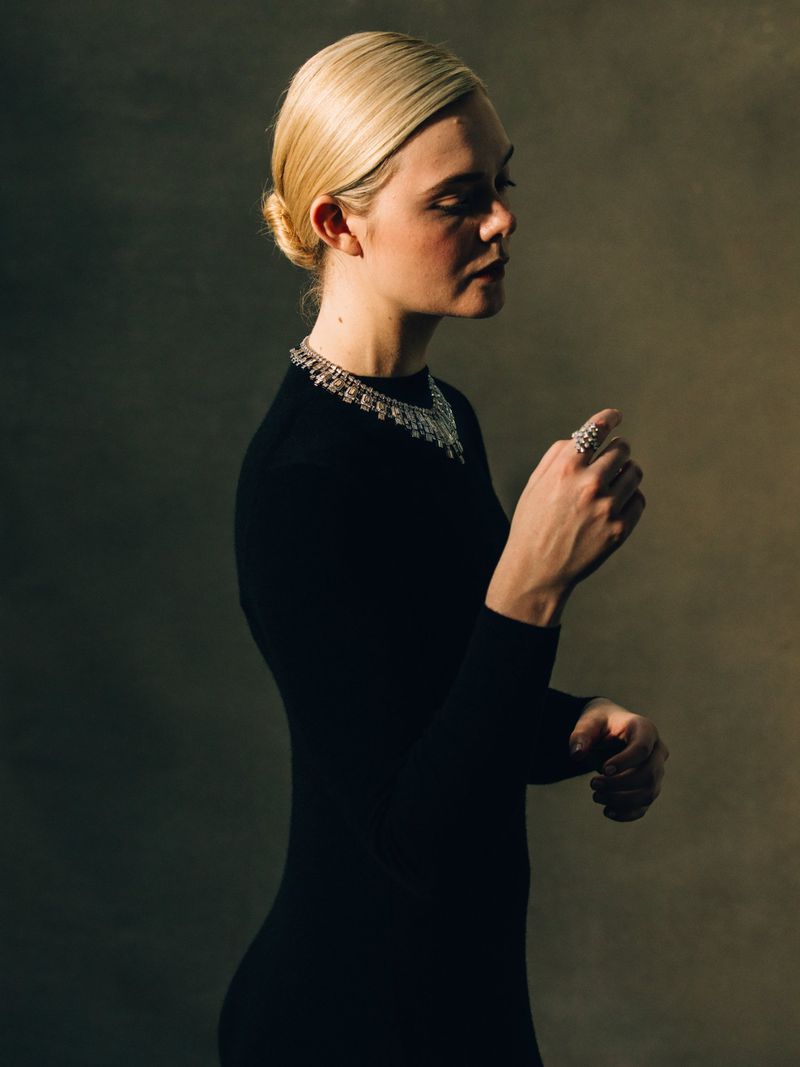 Elle Fanning in a black long-sleeve dress wearing a diamond Cartier necklace and ring against a muted backdrop.