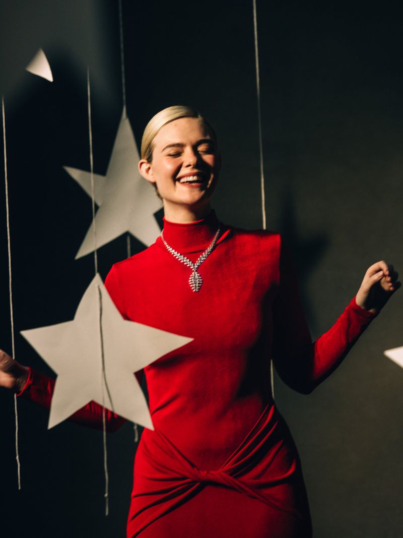Elle Fanning in a red high-neck dress and sparkling diamond necklace laughing amid hanging white star decorations.