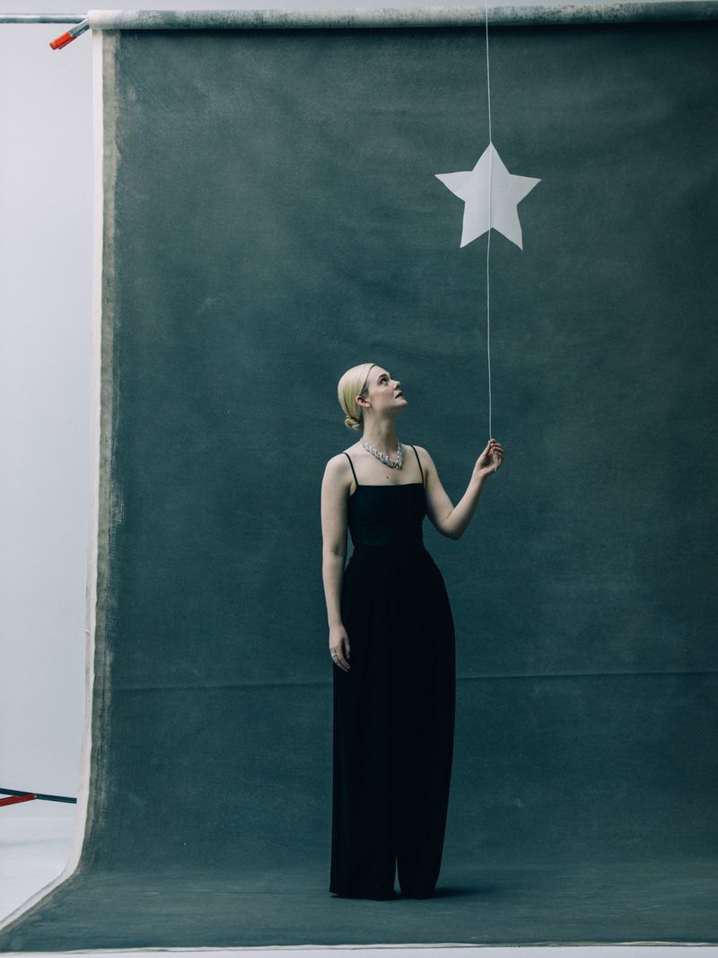 Elle Fanning wearing a black gown and sparkling necklace, looking up while holding a hanging white star prop against a textured studio backdrop.