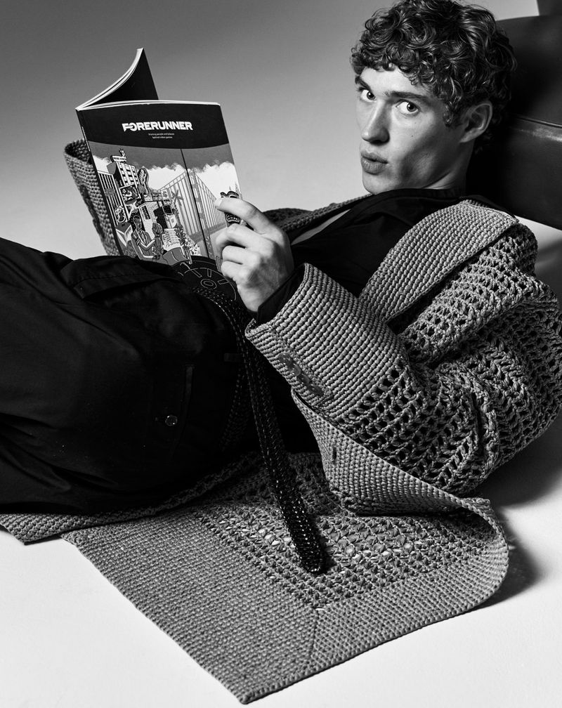 Black-and-white portrait of a young man lying down while reading a magazine titled 'Forerunner,' wearing a crocheted cardigan.