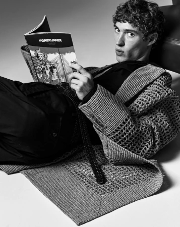 Black-and-white portrait of a young man lying down while reading a magazine titled 'Forerunner,' wearing a crocheted cardigan.
