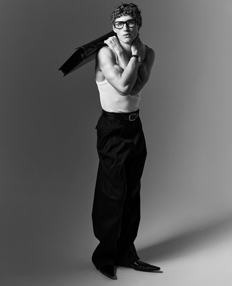 Valentin Humbroich poses in a white tank top, wide-leg black trousers, and glasses, holding a black folder over his shoulder.
