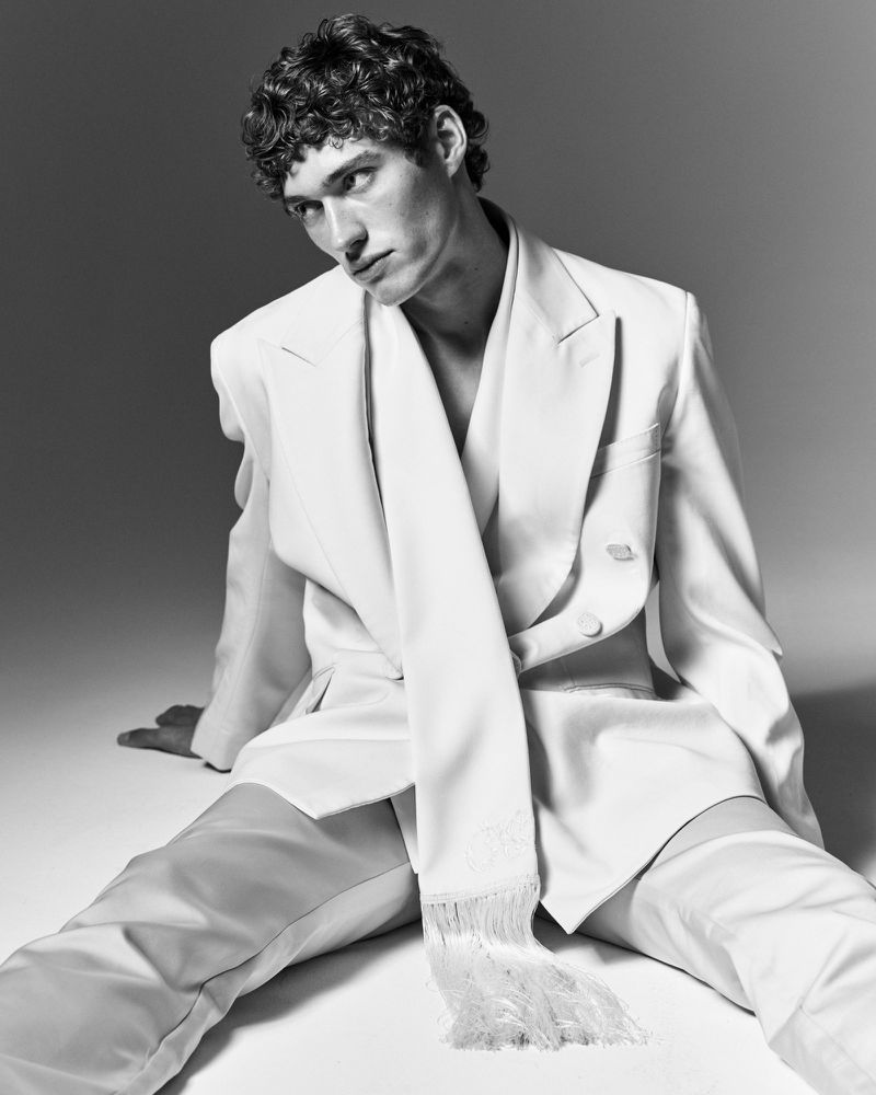 Monochrome portrait of a male model with curly hair wearing an off-white suit with a long scarf, posed in a seated position.
