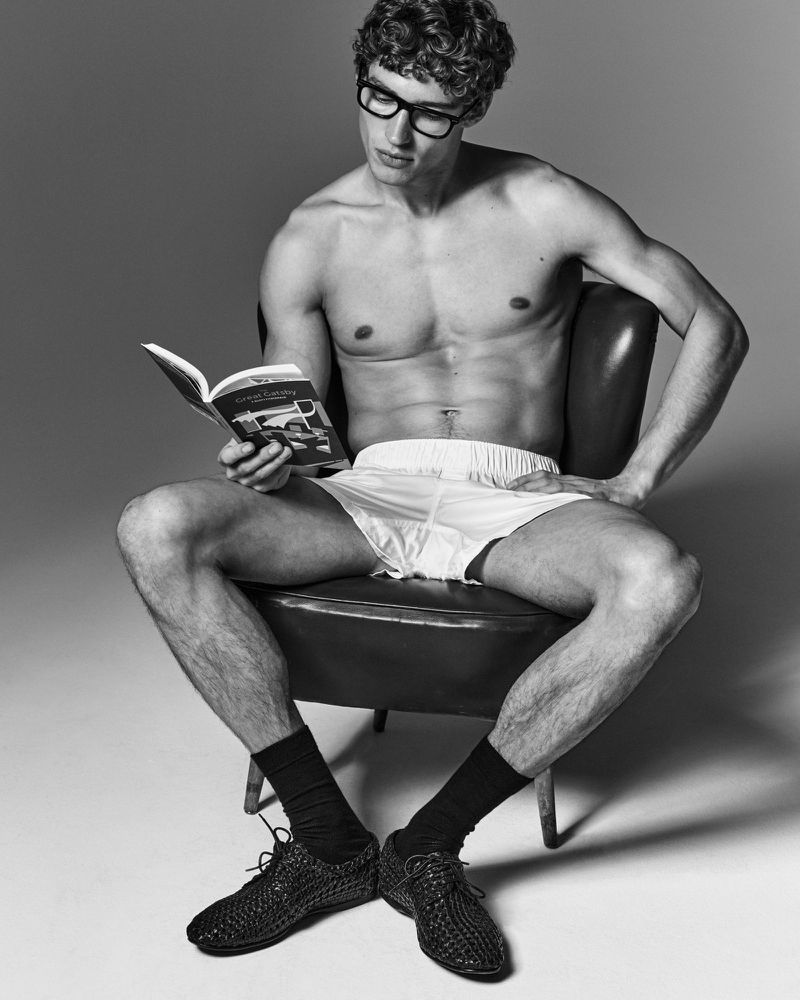 Black and white portrait of an adult male with glasses sitting in a chair, reading a book, wearing socks and shoes.