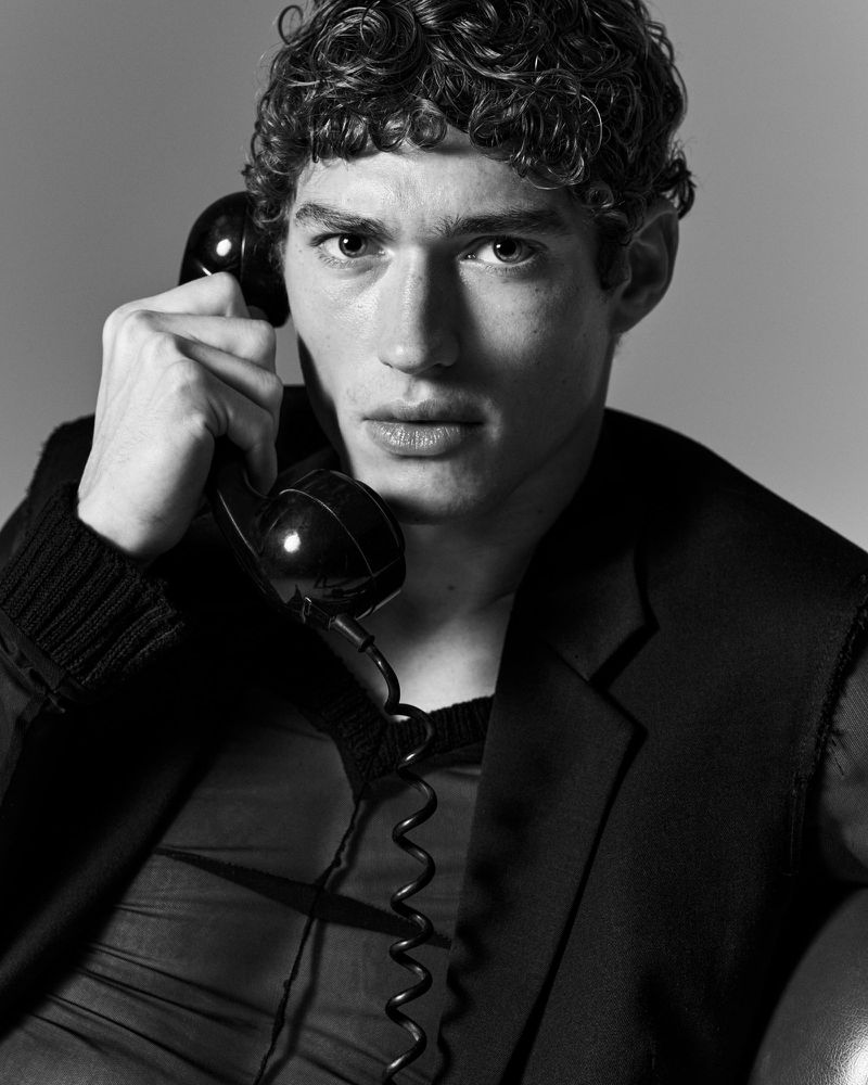 Black and white portrait of a male model with curly hair holding a rotary phone receiver to his ear in a studio setting.