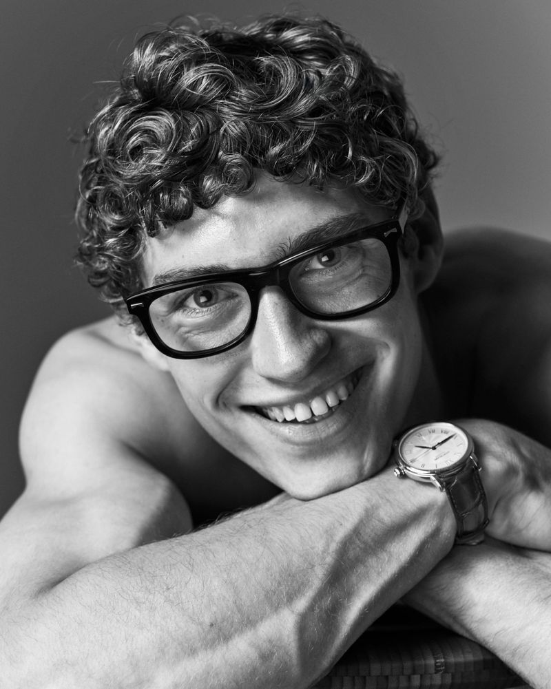Portrait of a smiling male model wearing black eyeglasses and a wristwatch, resting his chin on his arms in a studio setting.