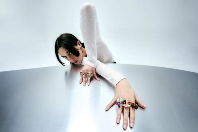 Model wearing multiple statement gemstone rings leaning over a curved metallic surface in a white studio