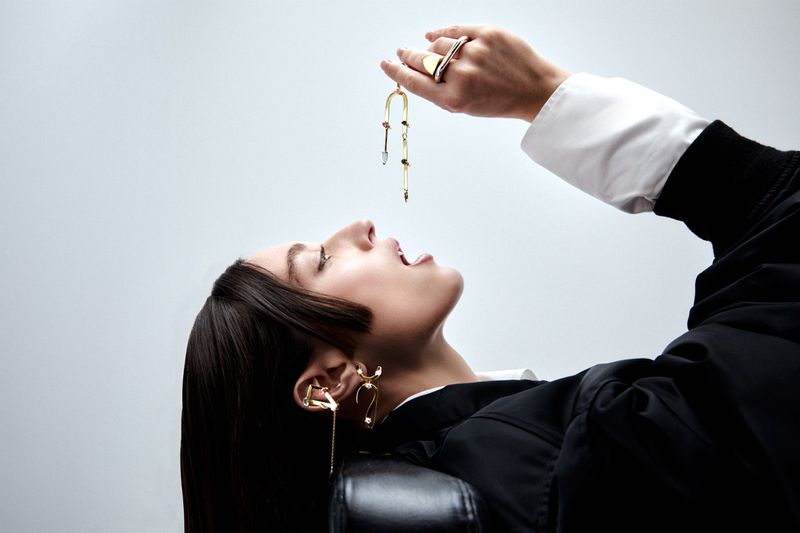 A model reclining with open mouth dangling a sculptural gold jewelry piece overhead against a seamless grey studio background.
