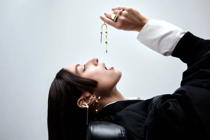 A model reclining with open mouth dangling a sculptural gold jewelry piece overhead against a seamless grey studio background.