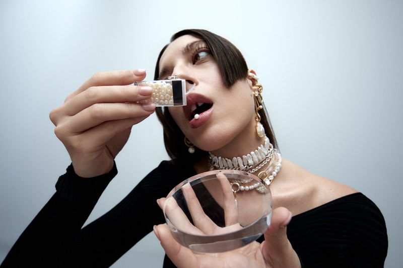 Close-up of a woman wearing pearl jewellery holding a clear container of pearls near her lip alongside a round acrylic disc.