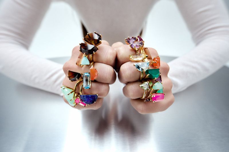 Two clenched fists adorned with multiple colorful gemstone rings resting on a reflective surface against a white backdrop.