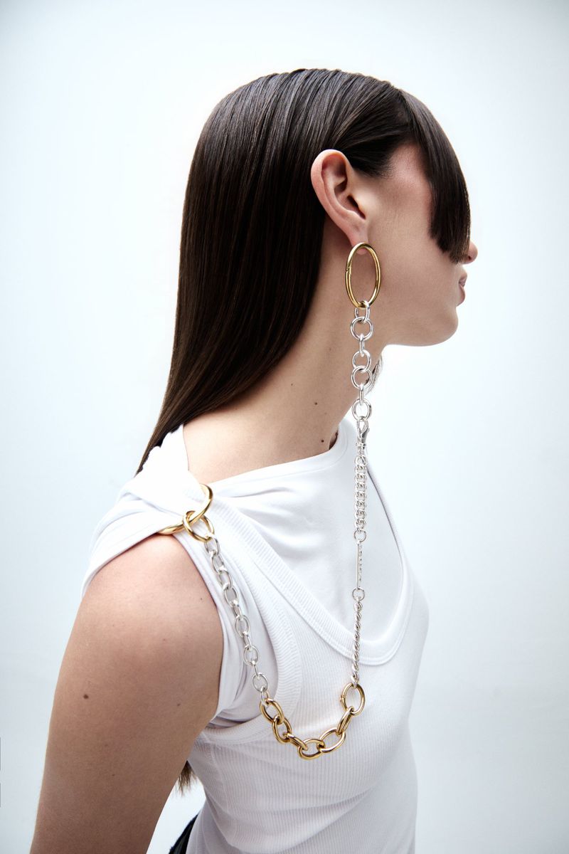Model in profile wearing a white tank top and a long mixed-metal chain earring connected to the shoulder against a white backdrop.