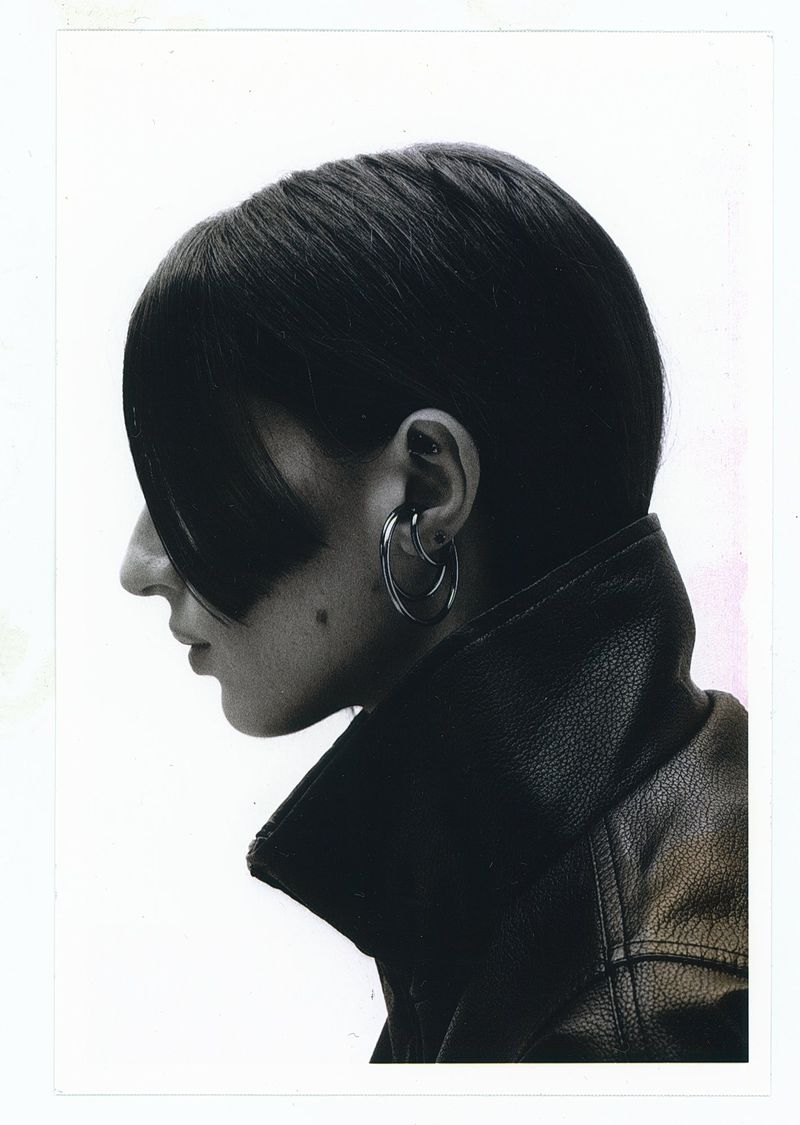 Profile portrait of a person with sleek dark fringe wearing sculptural silver hoop earrings and a high-collared leather jacket.