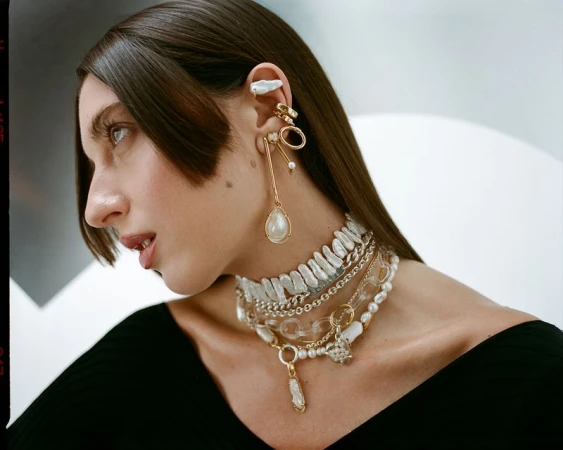 Close-up profile of a woman wearing layered pearl necklaces, gold chain chokers, and multi-piece ear cuffs and drop earrings.