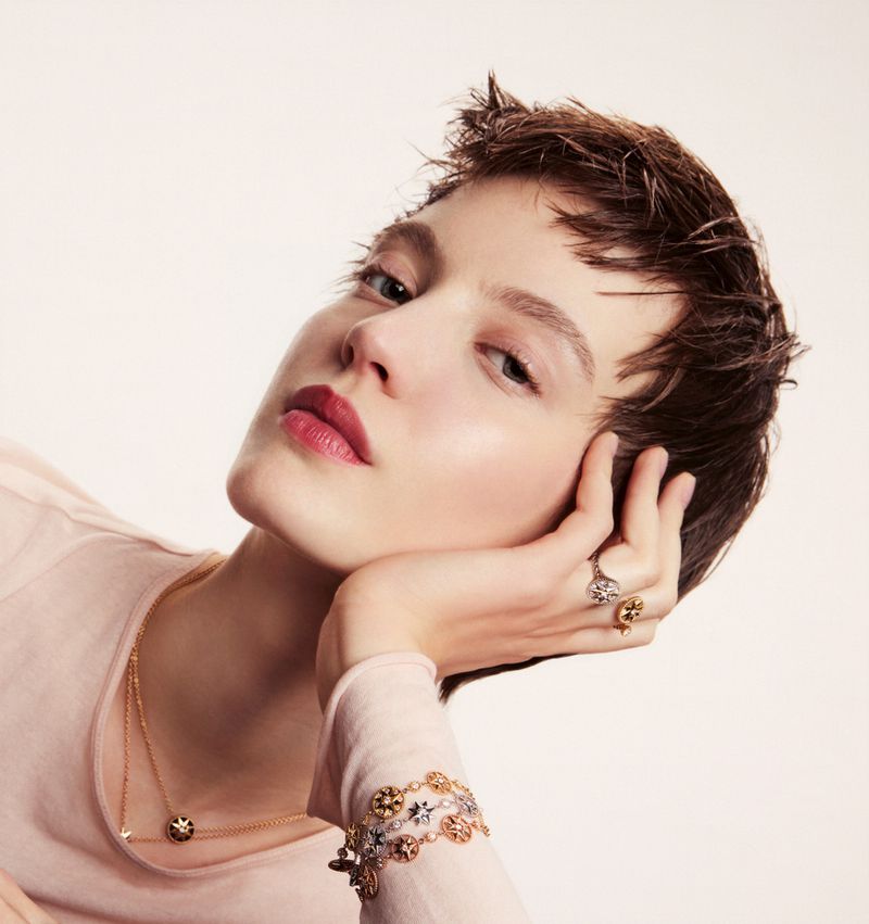Close-up portrait of a woman with short dark hair wearing delicate gold jewellery and rings against a plain light background