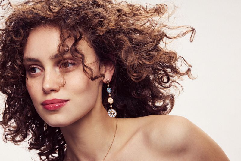 Close-up portrait of a woman with voluminous curly brown hair and dangling earrings against a white background.
