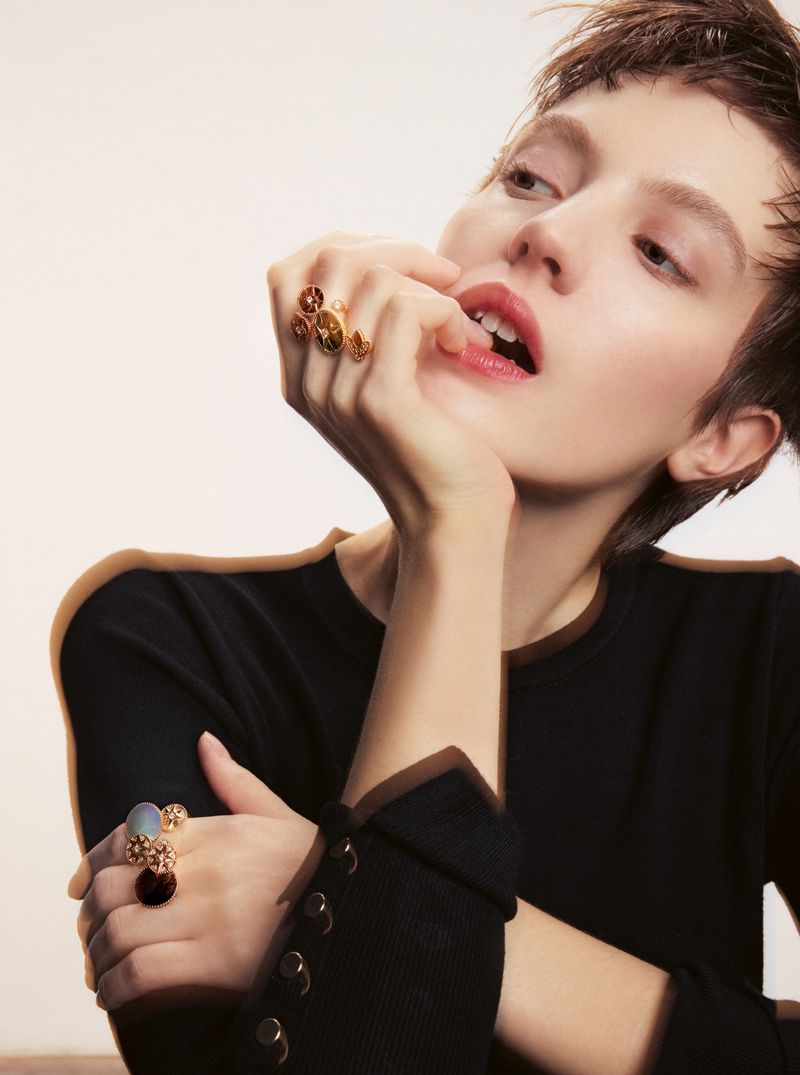 Model with short dark pixie hair wearing statement rings on both hands posing in a black top against a pale studio background.