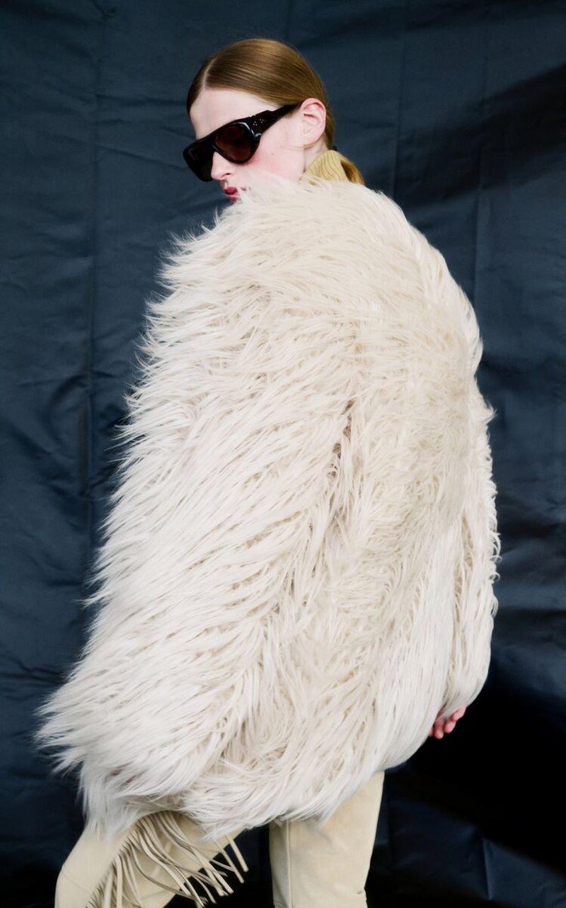 Model wearing a voluminous shaggy cream faux-fur coat and dark shield sunglasses against a dark textured backdrop.