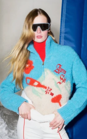Model wearing a blue après-ski fleece sweater, red turtleneck, white ski pants, and dark shield sunglasses.