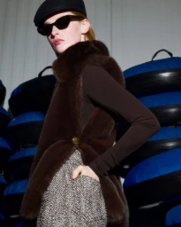 Model wearing dark sunglasses, a black cap, a plush brown fur vest, and tweed trousers against stacked blue snow tubes.