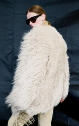 Model wearing a voluminous shaggy cream faux-fur coat and dark shield sunglasses against a dark textured backdrop.