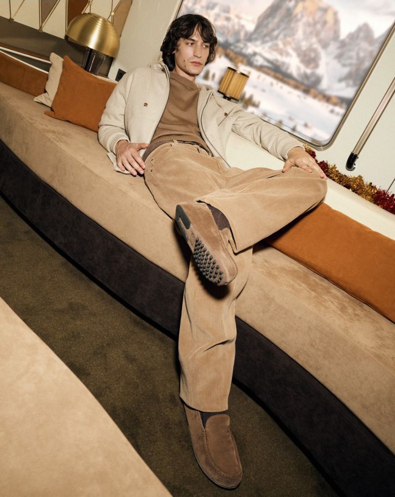 Valerio Maccario wearing beige corduroy trousers, cream jacket, and brown suede loafers while lounging on a plush bench.