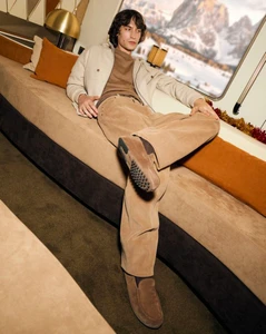 Valerio Maccario wearing beige corduroy trousers, cream jacket, and brown suede loafers while lounging on a plush bench.