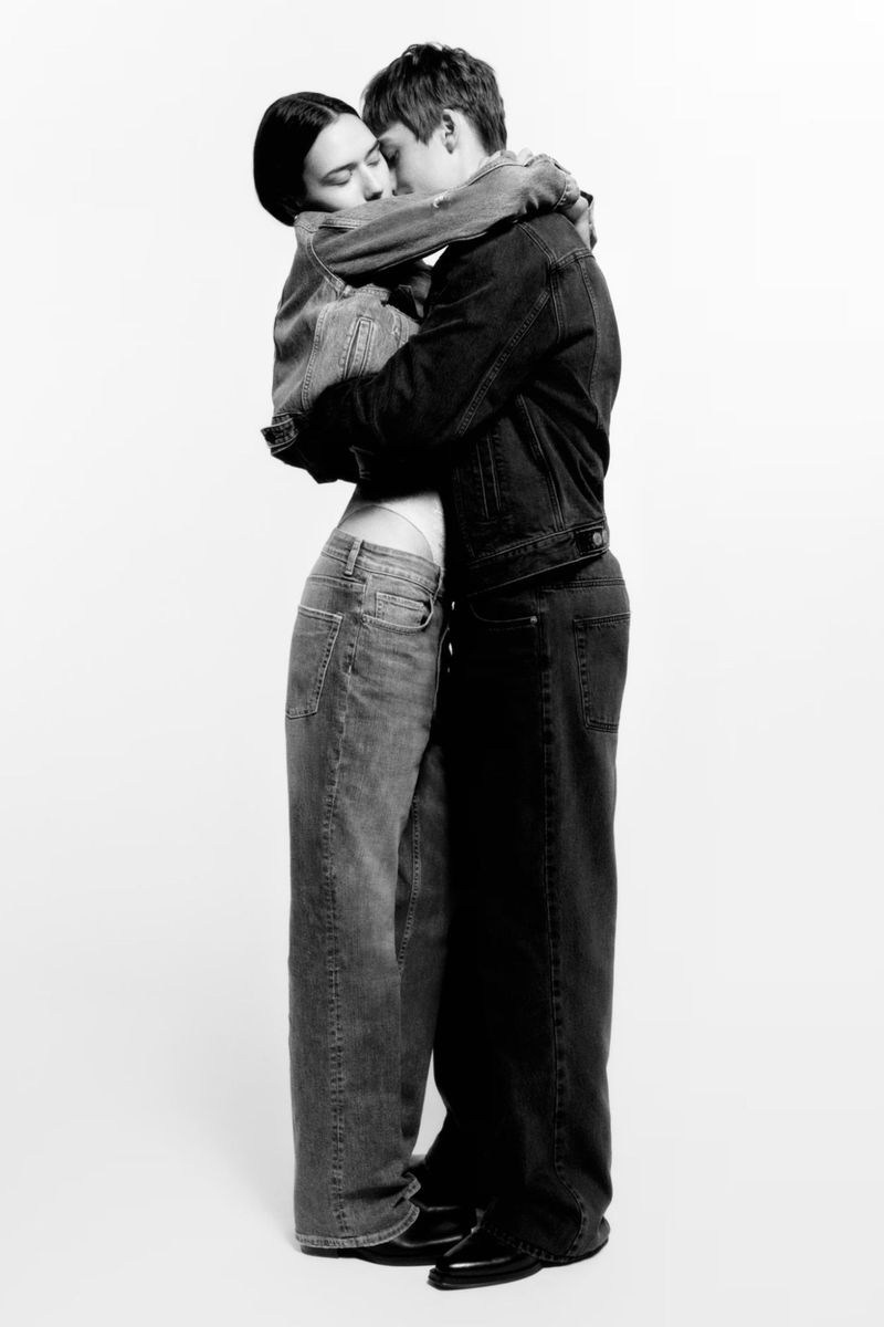 A couple embracing closely while wearing casual denim jackets and jeans against a bright white studio backdrop.