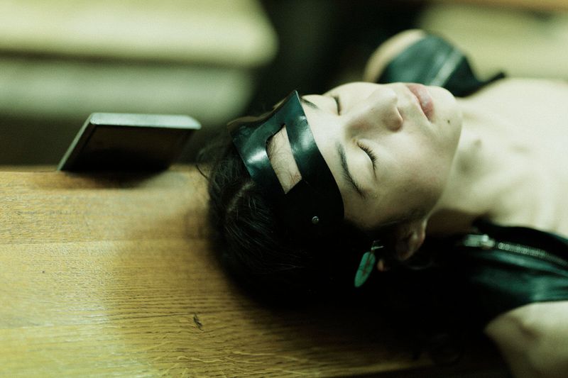 Model resting with eyes closed wearing a black cutout headpiece on a wooden surface backstage.
