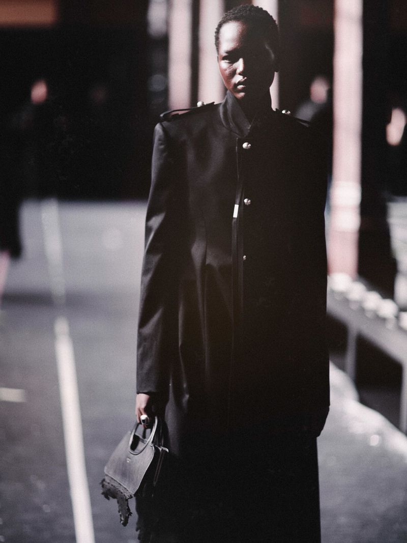 Model in a black military-style coat holding a handbag backstage at Ann Demeulemeester
