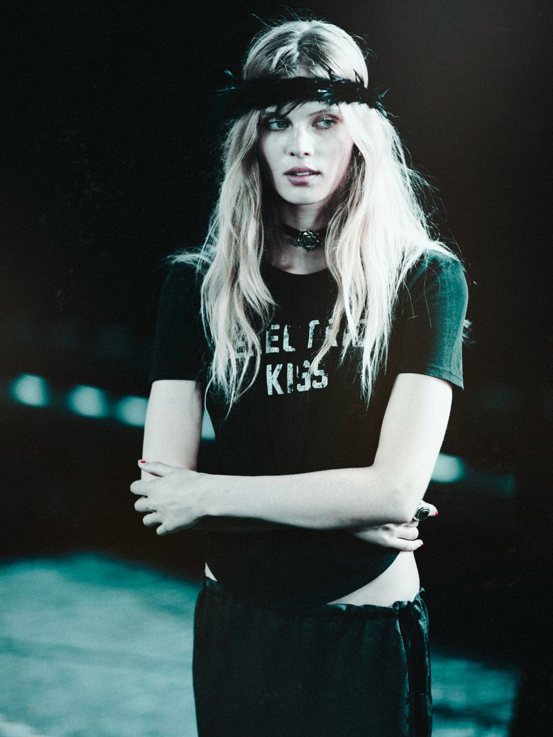 Blonde model wearing a feathered black headband, choker necklace, and black graphic t-shirt with crossed arms backstage.