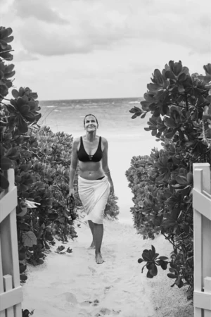 Black and white shot of a woman in a bikini and a white wrap walking on a sandy path towards the ocean between lush greenery.