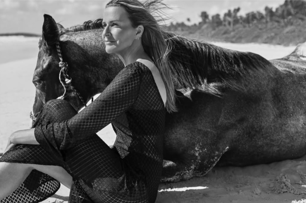 A woman in a patterned dress sits on a beach next to a horse. The image is black and white and the setting is a sunny day.