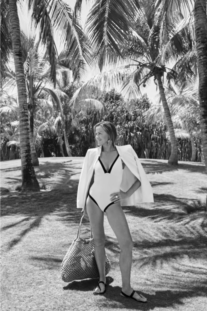 Woman in a swimsuit and blazer stands on a lawn surrounded by palm trees, holding a tote bag, in a sunny, black and white outdoor shot.