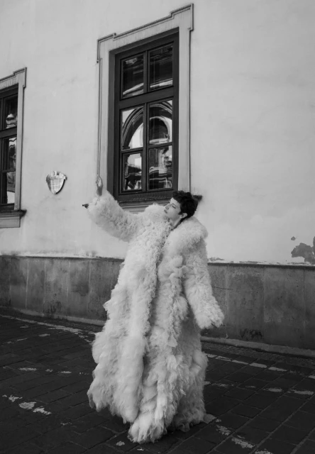 A woman in a fuzzy white coat stands on a brick sidewalk in front of a light-colored building with dark windows on a sunny day.