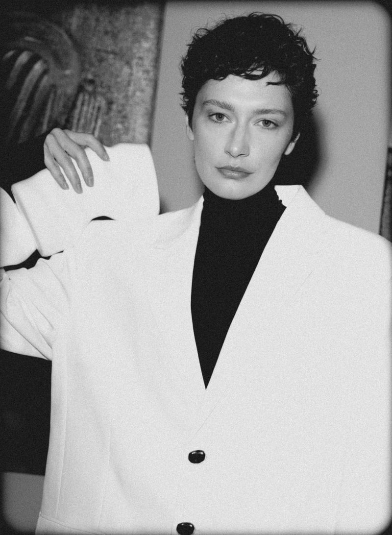 Black-and-white portrait of a female adult with short dark curly hair, wearing a black turtleneck and oversized white blazer, indoor.