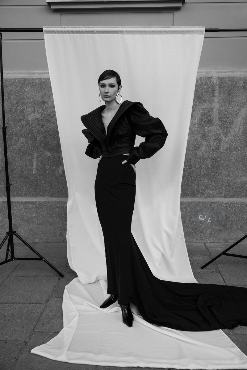 A woman in an elegant black gown poses in front of a white backdrop, showcasing high-fashion style in a monochromatic outdoor setting.