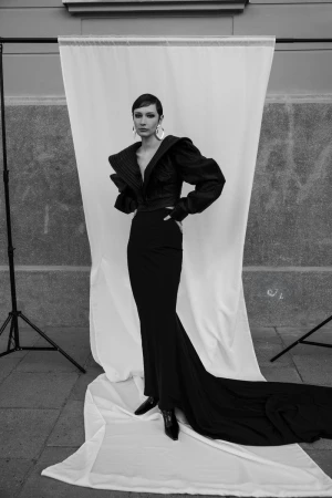 A woman in an elegant black gown poses in front of a white backdrop, showcasing high-fashion style in a monochromatic outdoor setting.