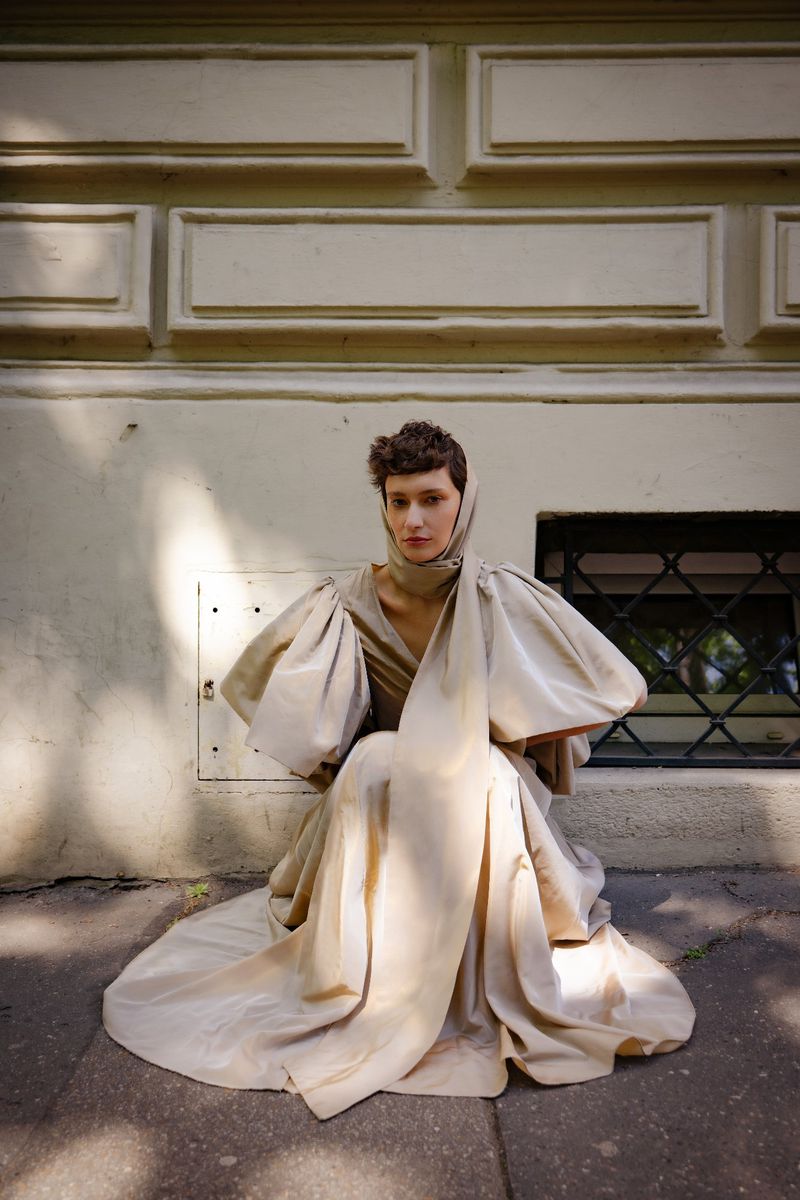 Woman poses in a dramatic beige dress with large balloon sleeves while kneeling on a stone surface against a cream coloured building wall.