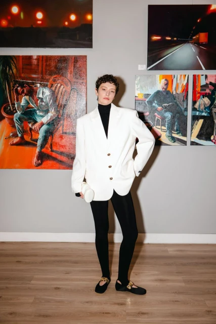 Woman in a white blazer poses in an art gallery in front of artwork. She's wearing black leggings and ballet flats with gold accents.