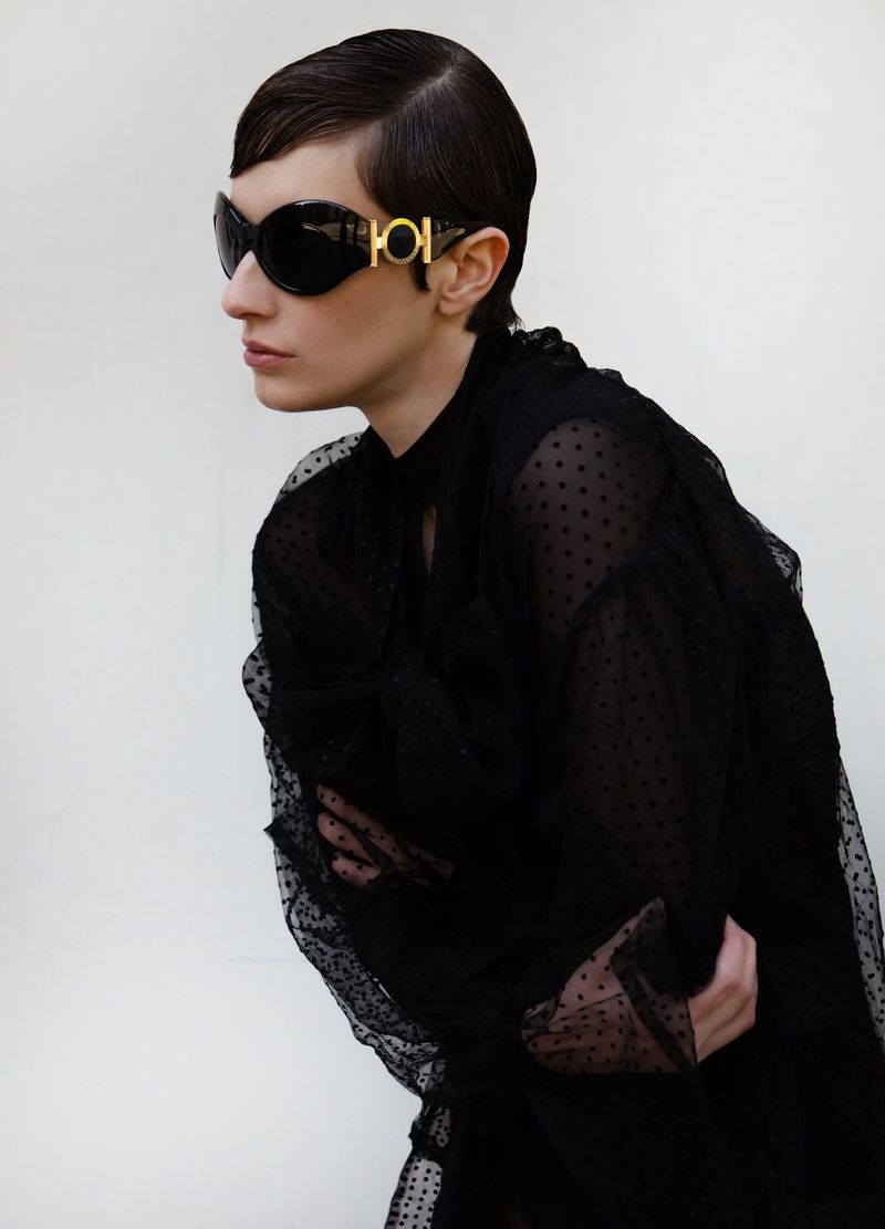 Woman poses in sheer black polka dot top and sunglasses with gold detail against a white backdrop in an eye-level portrait.