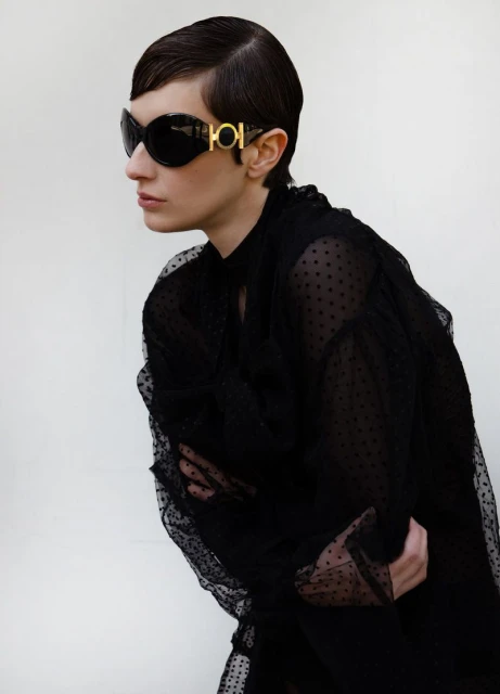 Woman poses in sheer black polka dot top and sunglasses with gold detail against a white backdrop in an eye-level portrait.